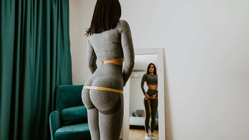smiling woman measuring body with tape standing in front of mirror